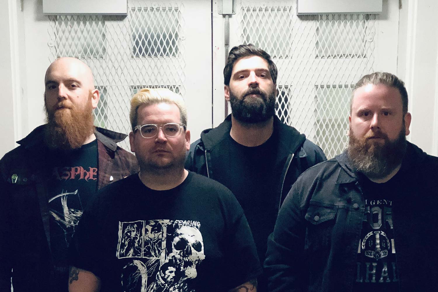 four dudes with beards and black tee shirts standing against steel doors