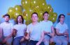 members of Dot.s sitting wearing white t-shirts with yellow balloons in the background.