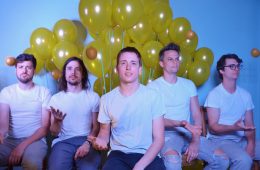 members of Dot.s sitting wearing white t-shirts with yellow balloons in the background.