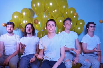 members of Dot.s sitting wearing white t-shirts with yellow balloons in the background.