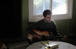 Alex Lotito playing an acoustic guitar and writing lyrics at a kitchen table
